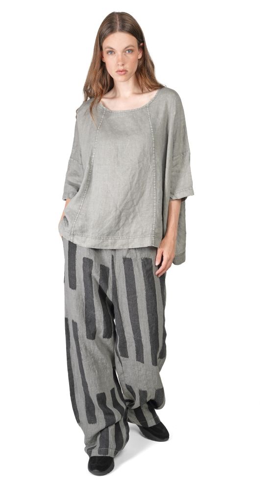 Frau trägt oversized graues Hemd mit weitem Schnitt und gestreifte, weite Hosen, barfuß auf hellem Hintergrund.