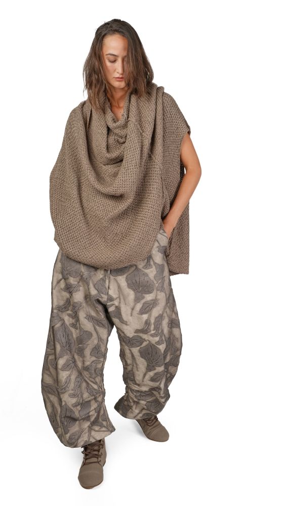 Person trägt oversized Strickbluse mit weitem Rock und gemusterten Hosen, barfuß auf hellem Hintergrund.