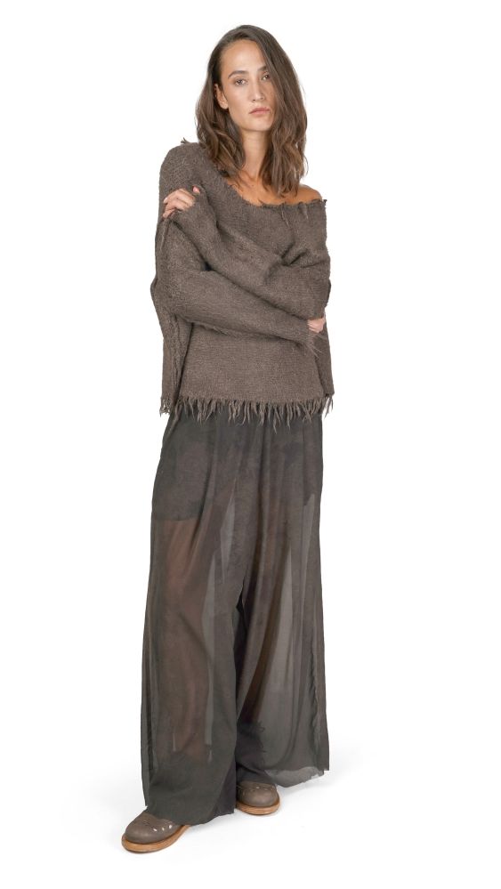 Frau trägt oversized grauen Pullover mit Fransen, dunkle weite Hose und flache Schuhe, Arme verschränkt.