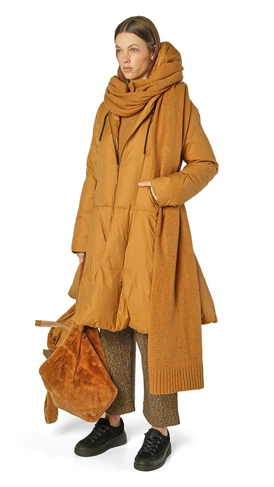Frau trägt langen, warmen camelfarbenen Mantel mit Schal und hält braune Tasche, schwarze Schuhe.