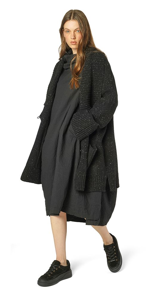 Frau trägt oversized schwarzen Strickmantel mit Knit-Einsätzen und schwarze Sneakers.