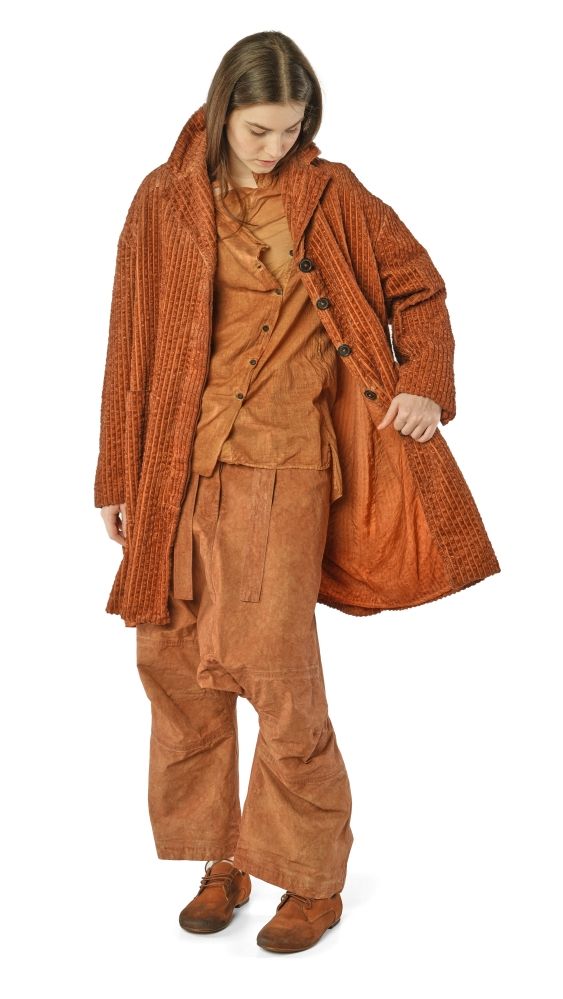 Frau trägt rustikale, braune Wollkleidung mit Cape und weitem Hosenrock, hält Tasche.
