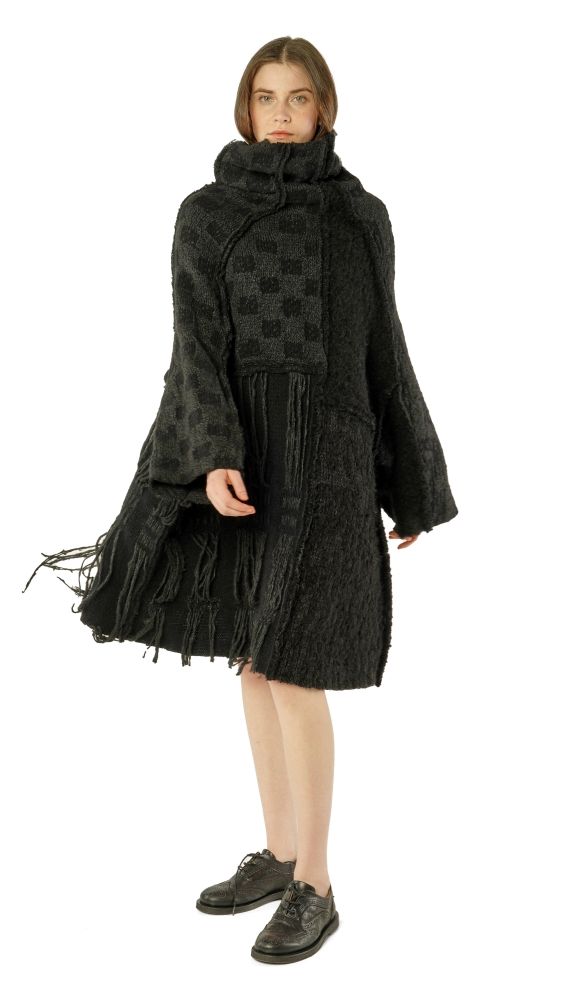 Frau trägt oversized schwarzen Wollmantel mit Fransen und kurzem Rock, barfuß mit schwarzen Sneakern.
