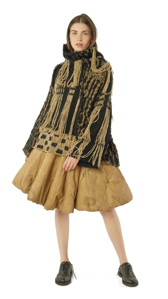 Frau trägt oversized gestrickte Cape-Jacke mit Fransen, beige weiten Rock und schwarze Stiefel.