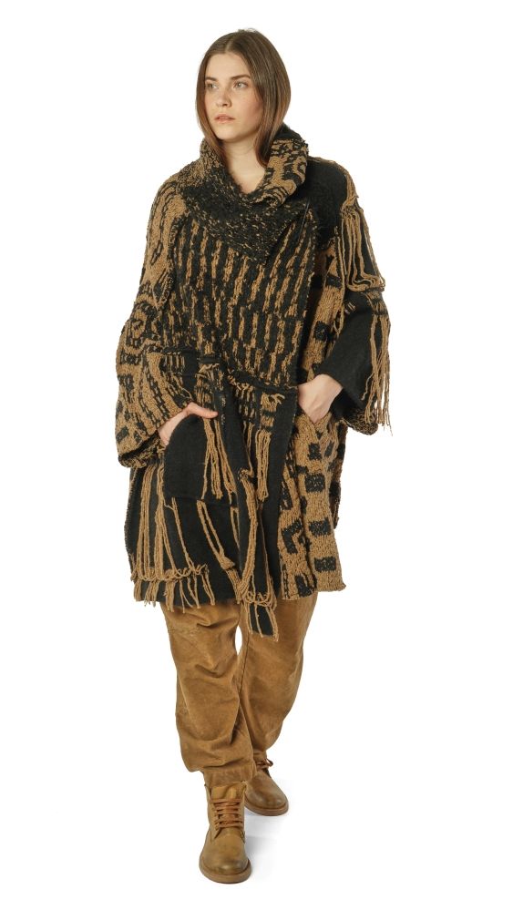 Frau trägt oversized gestrickten Poncho mit Fransen in Schwarz-Gold, dazu beige Hosen und Stiefel.