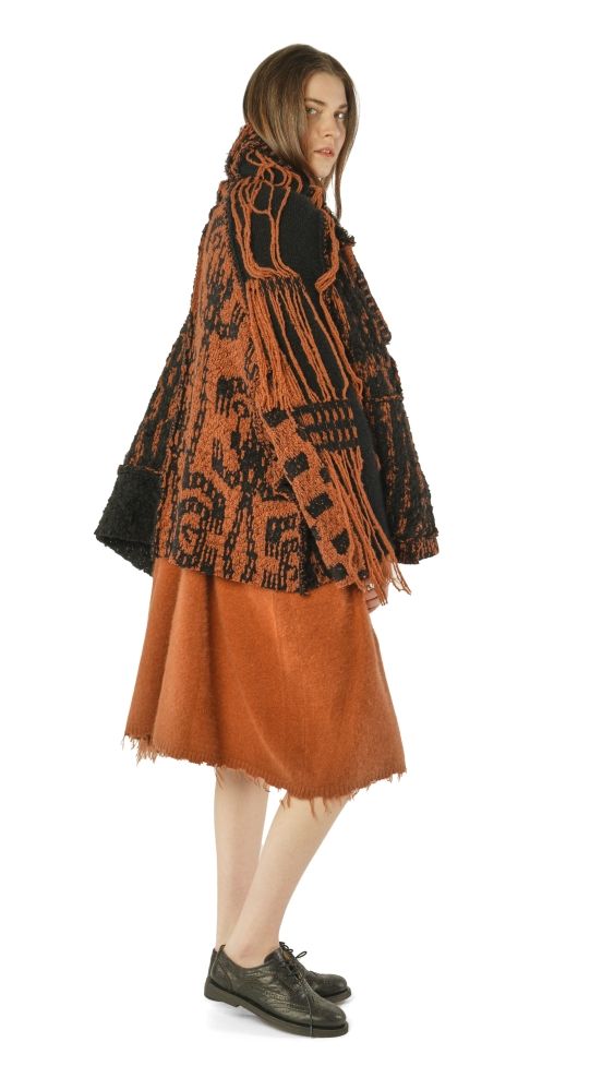 Person trägt buntes, gemustertes Cape mit orangefarbenem Rock und schwarzen Schuhen.