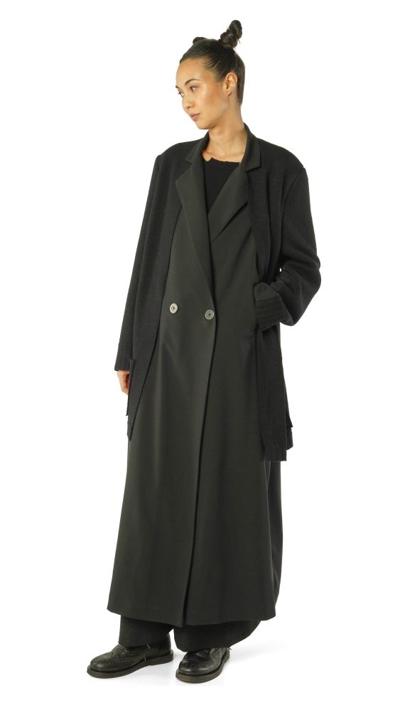 Schlanke Person in langem schwarzem Trenchcoat mit schwarzen Stiefeln und dunkler Kleidung.