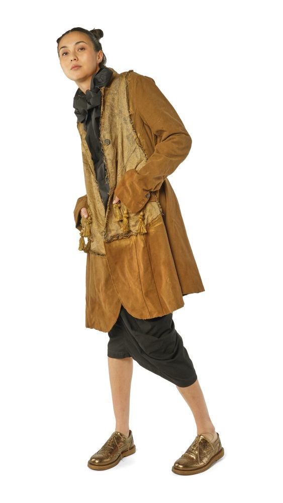 Person trägt langen, braunen Mantel mit Muster, schwarze Hose und goldene Schuhe, hält Tasche.