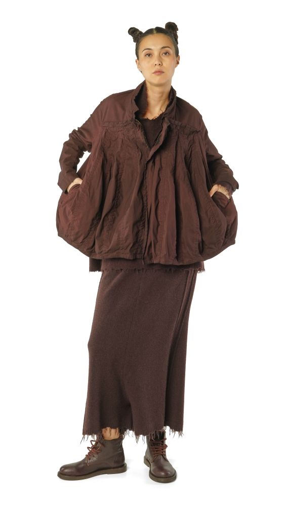 Person trägt oversized braunen Mantel mit weitem Rock und robusten Stiefeln, Haare zu zwei Knoten gebunden.
